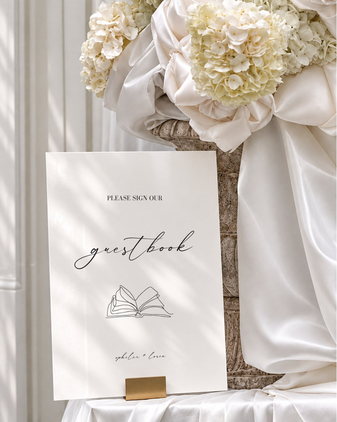 Aria acrylic wedding guestbook sign in a gold holder beside draped ivory silk and hydrangeas