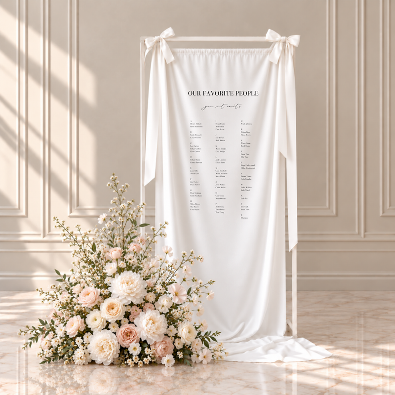 Aria fabric wedding seating chart: our favorite people, your seat awaits, with guests listed A to Z in three columns, hanging from a white frame with ribbon bows beside blush and ivory flowers