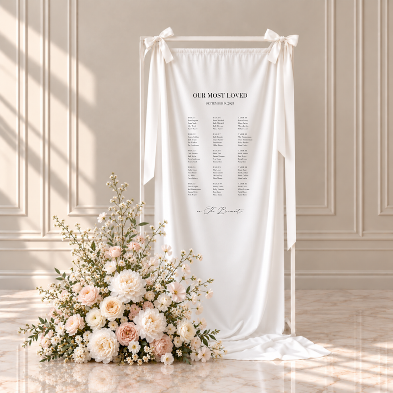 Aria fabric wedding seating chart: our most loved, with guests grouped by table across fifteen tables and a family signature, hanging from a white frame with ribbon bows beside blush and ivory flowers