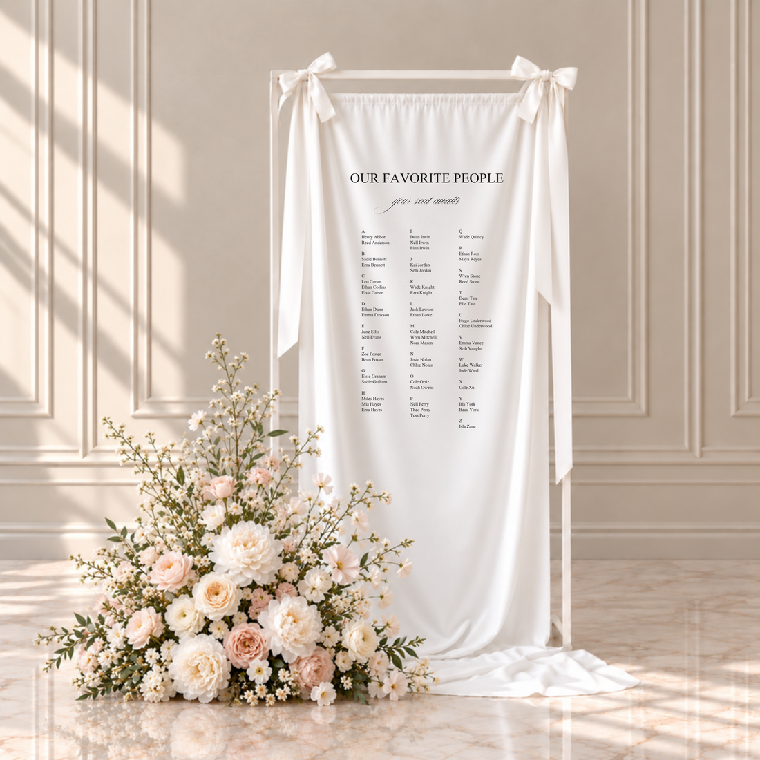Florence fabric wedding seating chart: our favorite people, your seat awaits, with guests listed A to Z in three columns, hanging from a white frame with ribbon bows beside blush and ivory flowers