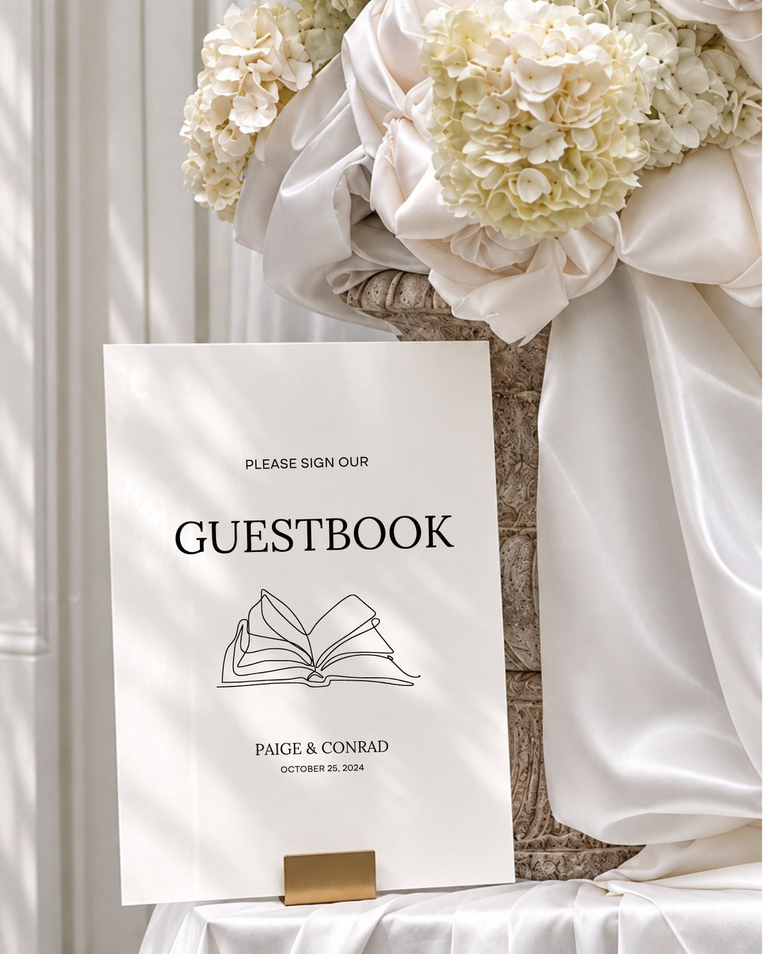 Moda acrylic wedding guestbook sign in a gold holder beside draped ivory silk and hydrangeas