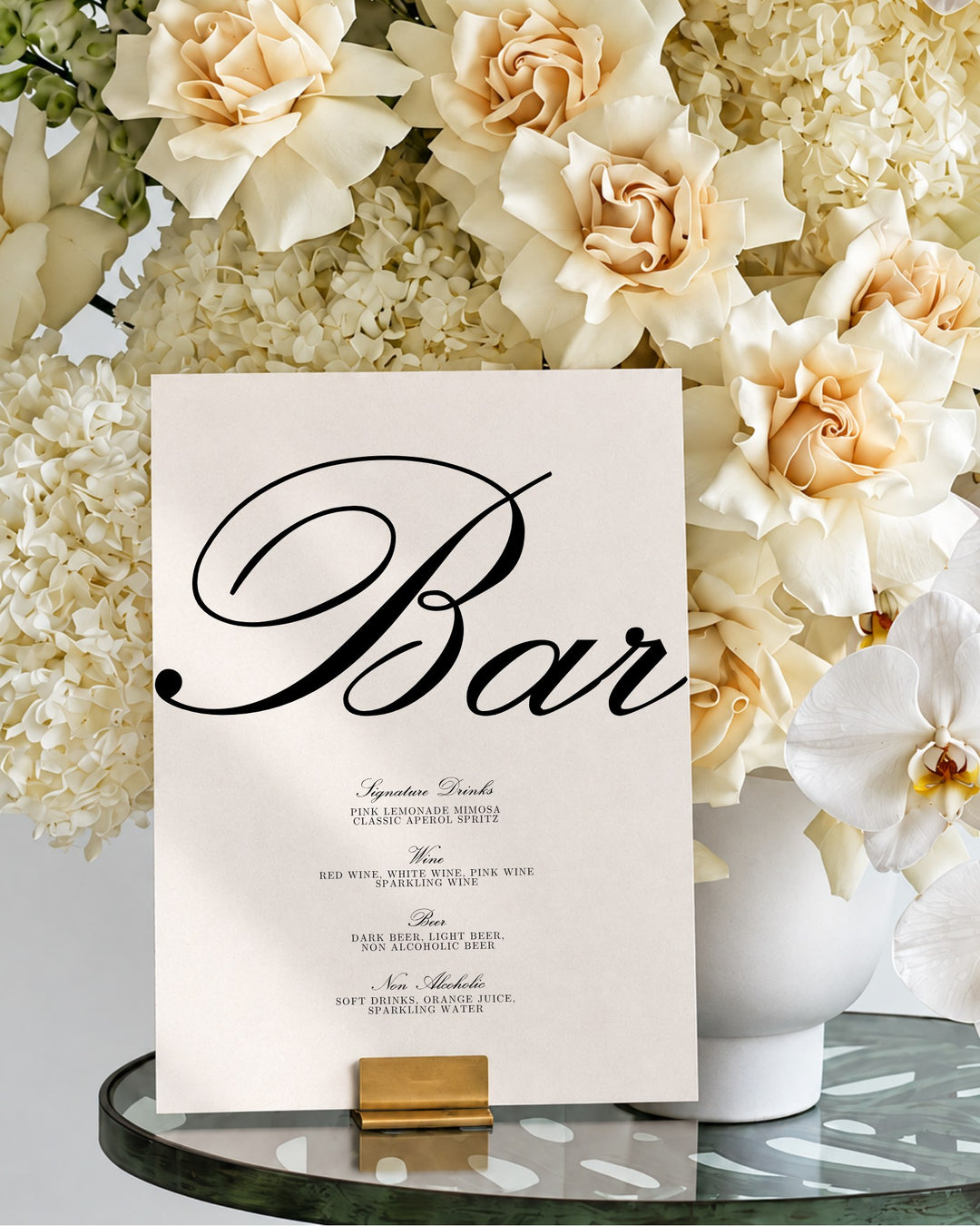 Reina acrylic wedding bar and drinks sign in a gold holder on a glass table in front of ivory roses and hydrangeas